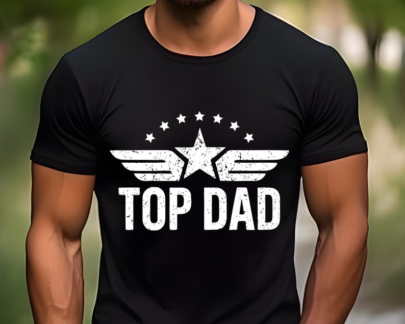 K&ouml;nnte beinhalten: Schwarzes Rundhals-T-Shirt mit einem wei&szlig;en, verwitterten Aufdruck. Der Aufdruck zeigt einen Stern, Fl&uuml;gel und den Text "TOP DAD" in einer fetten, serifenlosen Schrift. Das Design ist auf der Brust zentriert.