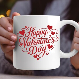 May include: White ceramic mug with a red "Happy Valentine's Day" design. The design includes hearts and decorative swirls. The mug has a curved handle and is held by a person.