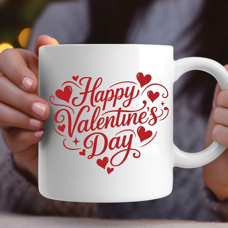 May include: White ceramic mug with a red "Happy Valentine's Day" design. The design includes hearts and decorative swirls. The mug has a curved handle and is held by a person.
