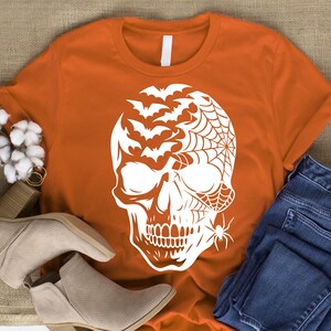 May include: An orange t-shirt with a white skull design, incorporating bats, a spider web, and a spider. This Halloween-themed tee is made from a soft fabric, perfect for spooky occasions.