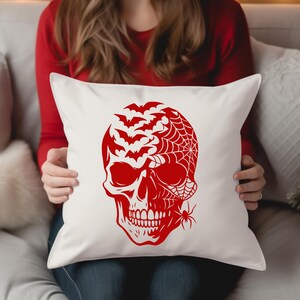 May include: Square white cushion with a red Halloween design. The design features a skull with bats, a spider web, and a spider. The cushion is held by a person wearing a red long-sleeve top.