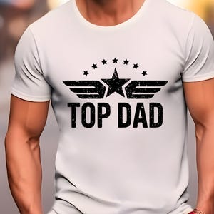 K&ouml;nnte beinhalten: Wei&szlig;es T-Shirt mit schwarzem Aufdruck. Der Aufdruck zeigt einen Stern, Fl&uuml;gel und den Text "TOP DAD". Das Design ist mittig auf der Brust platziert. Das Shirt hat kurze &Auml;rmel und einen Rundhalsausschnitt.
