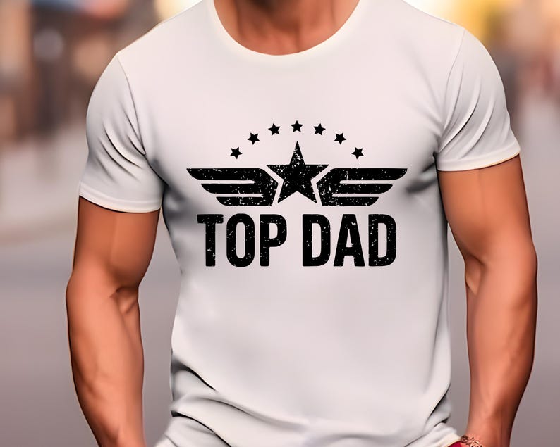 K&ouml;nnte beinhalten: Wei&szlig;es T-Shirt mit schwarzem Aufdruck. Der Aufdruck zeigt einen Stern, Fl&uuml;gel und den Text "TOP DAD". Das Design ist mittig auf der Brust platziert. Das Shirt hat kurze &Auml;rmel und einen Rundhalsausschnitt.