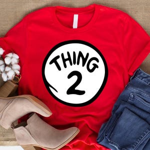 May include: A red t-shirt featuring a white circle with the text "THING 2" in black. The shirt is paired with blue jeans and beige ankle boots. The design is a playful nod to a popular children's book.