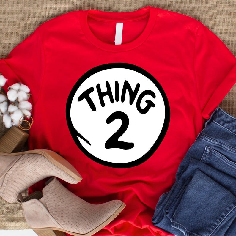 May include: A red t-shirt featuring a white circle with the text "THING 2" in black. The shirt is paired with blue jeans and beige ankle boots. The design is a playful nod to a popular children's book.