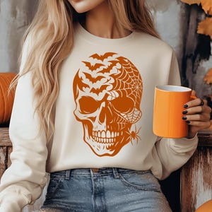 May include: Cream-coloured sweatshirt with an orange Halloween skull design, including bats, a spiderweb, and a spider. An orange mug is held in the hand. Ideal for the spooky season.