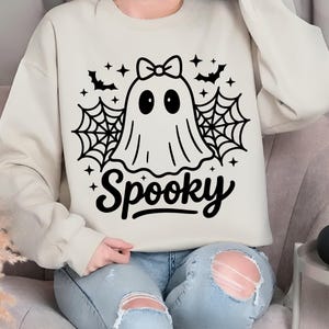 May include: Cream-colored sweatshirt with a black graphic of a ghost wearing a bow, surrounded by spiderwebs, bats, and stars. The word "Spooky" is written in a script font below the ghost. A casual, festive garment for Halloween.