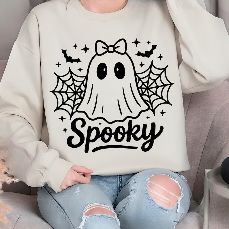 May include: Cream-colored sweatshirt with a black graphic of a ghost wearing a bow, surrounded by spiderwebs, bats, and stars. The word "Spooky" is written in a script font below the ghost. A casual, festive garment for Halloween.
