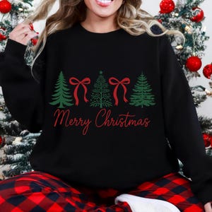 以下が含まれることがあります： 緑のクリスマスツリーのデザインと赤いリボンが付いた黒いスウェットシャツ。「Merry Christmas」の文字が木の下に赤い文字で書かれています。画像内の人物は、赤と黒のチェック柄のパジャマパンツを着用しています。