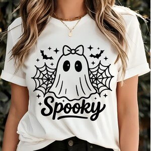 May include: White t-shirt with a black graphic of a ghost wearing a bow, surrounded by bats, spiderwebs, and stars. The word "Spooky" is written in a script font below the ghost. The shirt has rolled-up sleeves.