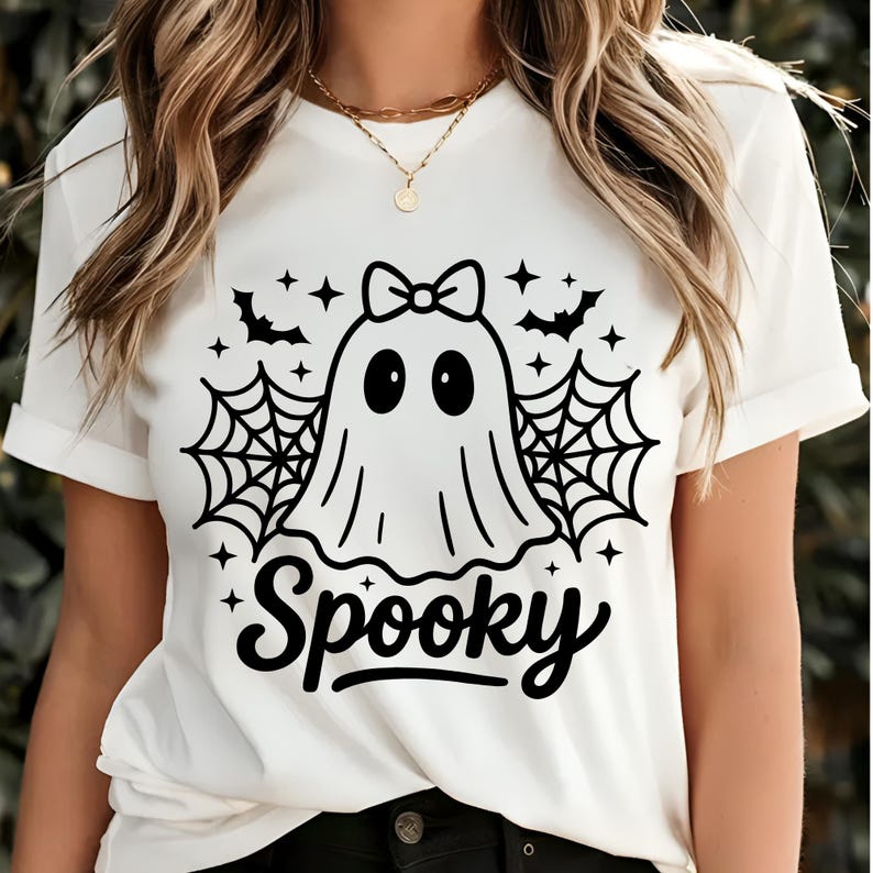 May include: White t-shirt with a black graphic of a ghost wearing a bow, surrounded by bats, spiderwebs, and stars. The word "Spooky" is written in a script font below the ghost. The shirt has rolled-up sleeves.