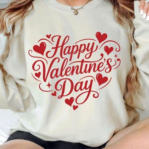 May include: Cream-colored sweatshirt with a red "Happy Valentine's Day" design. The design features the text in a heart shape, with red hearts and decorative swirls. A festive and romantic apparel item.