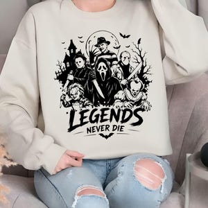 May include: A beige crewneck sweatshirt featuring a black graphic print of various horror movie characters, including a masked figure, a doll, and a clown, set against a haunted house and moon background with the text "LEGENDS NEVER DIE".