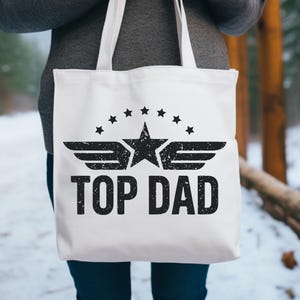 K&ouml;nnte beinhalten: Wei&szlig;e Tragetasche mit schwarzem Text und Grafik. Das Design zeigt einen Stern mit Fl&uuml;geln, umgeben von Sternen, und die Worte "TOP DAD". Die Tasche hat wei&szlig;e Griffe und wird von einer Person gehalten.