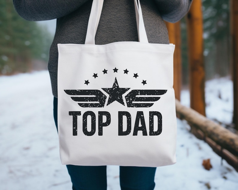 K&ouml;nnte beinhalten: Wei&szlig;e Tragetasche mit schwarzem Text und Grafik. Das Design zeigt einen Stern mit Fl&uuml;geln, umgeben von Sternen, und die Worte "TOP DAD". Die Tasche hat wei&szlig;e Griffe und wird von einer Person gehalten.