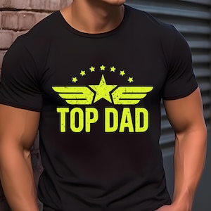 K&ouml;nnte beinhalten: Schwarzes T-Shirt mit neongelber Grafik. Die Grafik zeigt einen Stern, Fl&uuml;gel und den Text "TOP DAD". Das Design ist leicht verblichen. Das Shirt hat kurze &Auml;rmel und scheint aus einem weichen Material zu sein.