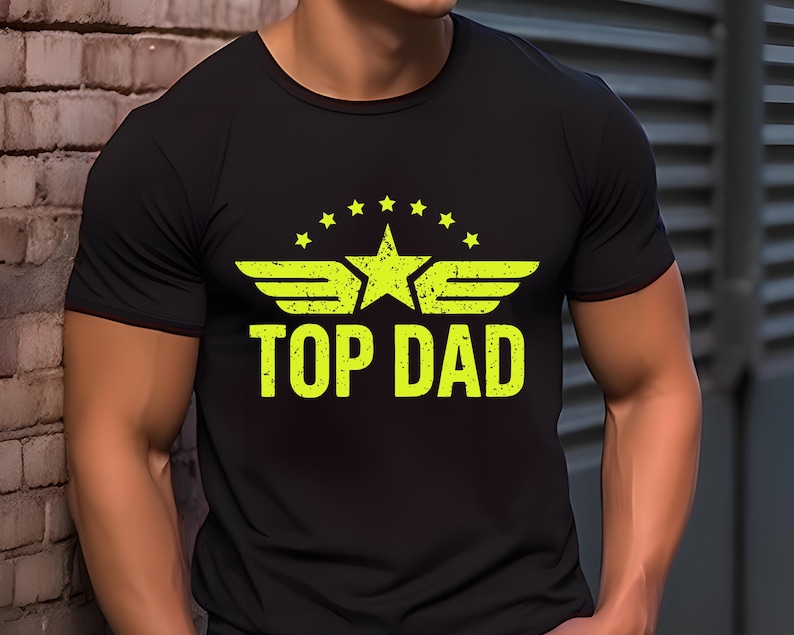 K&ouml;nnte beinhalten: Schwarzes T-Shirt mit neongelber Grafik. Die Grafik zeigt einen Stern, Fl&uuml;gel und den Text "TOP DAD". Das Design ist leicht verblichen. Das Shirt hat kurze &Auml;rmel und scheint aus einem weichen Material zu sein.