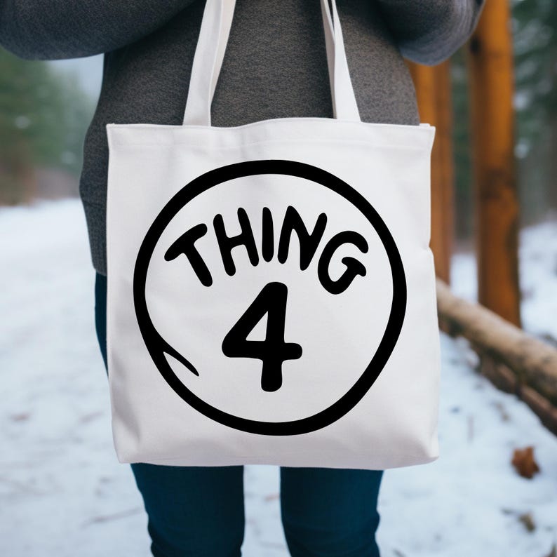 May include: White tote bag with a black circular design featuring the words "THING 4". The design is in a bold, playful font. The bag has white straps and is held by a person wearing a gray sweater and blue jeans.