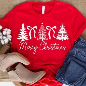 以下が含まれることがあります： クリスマスツリーとリボン、そして「Merry Christmas」の文字が白で描かれた赤いTシャツ。青いジーンズとベージュのアンクルブーツと組み合わせています。