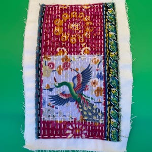 May include: A textile art piece with a white border and a central design featuring a peacock. The peacock is depicted with blue, green, and red hues, set against a background of floral patterns and a deep red base. The piece is hand-stitched.