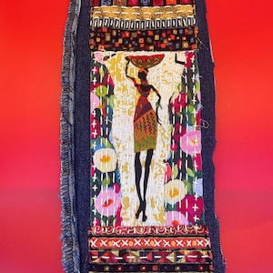 May include: A denim fabric art piece featuring an African woman carrying a basket on her head. The artwork incorporates colorful floral and geometric patterns, with a red background. The piece is framed with a denim border.