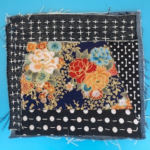 May include: A square fabric piece with a patchwork design. The top and left sides feature dark denim with white cross stitch patterns. The center showcases a floral print with red, orange, and blue flowers on a navy background. Black and white polka dot fabric is on the right and bottom.