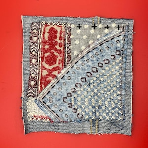 May include: A square textile art piece with a denim border and various fabric patches. The patches feature red, white, and blue patterns, including dots, geometric shapes, and floral designs. The piece is stitched together with white and black thread.