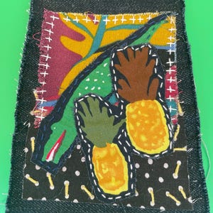 Pode incluir: Obra de arte têxtil com um jacaré verde e dois abacaxis amarelos. O desenho é costurado em tecido preto com bolinhas brancas e uma borda de ganga escura. A obra de arte tem uma estética artesanal e caprichosa.