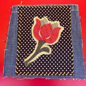 Puede incluir: Un parche de mezclilla con un diseño floral. El parche presenta una flor roja con detalles grises y amarillos sobre un fondo de lunares blancos y negros. La flor está cosida a la tela con hilo amarillo.