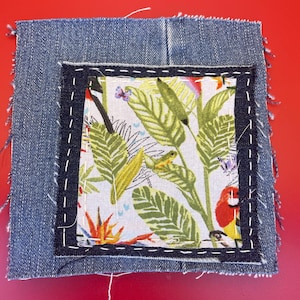 Op de afbeelding: Een vierkante denim patch met een vierkante stof met tropische print in het midden gestikt. De stof heeft groene bladeren, een papegaai en een kikker. De denim heeft een gerafelde rand en is gestikt met witte draad.