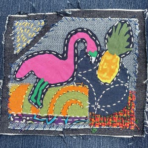 Pode incluir: Um patch de ganga com um flamingo rosa e um ananás. O flamingo tem um contorno preto e detalhes verdes. O ananás é amarelo e verde. O patch tem um fundo azul com detalhes de tecido coloridos e costuras brancas.