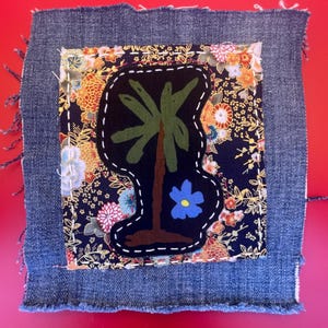 May include: A square denim patch with a fringed edge. The patch features a black palm tree design with a blue flower on a floral background. The palm tree is stitched with white thread.