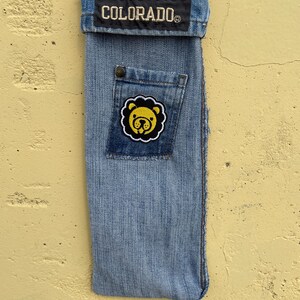 May include: A denim phone case with a stitched-on patch featuring a yellow lion's head. The case has a pocket and a strap. The word "COLORADO" is embroidered on a patch at the top.