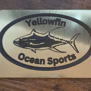 May include: A rectangular, brushed metal plaque with rounded corners. The plaque features the words "Yellowfin Ocean Sports" in black, surrounding a black illustration of a yellowfin tuna fish.