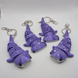 May include: Four articulated purple shark keychains with silver-toned metal rings. Two shark figures feature white dot patterns, while the other two display white constellation designs. Each shark has a wide, open mouth and dark eye details.
