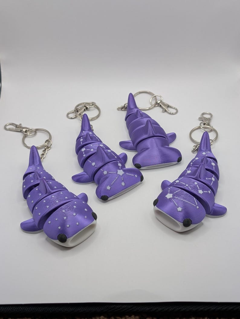 May include: Four articulated purple shark keychains with silver-toned metal rings. Two shark figures feature white dot patterns, while the other two display white constellation designs. Each shark has a wide, open mouth and dark eye details.