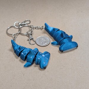 May include: Two blue hammerhead shark keychains with silver key rings and clasps. The shark keychains are segmented and have a speckled pattern. A US quarter is next to the keychains for size comparison.