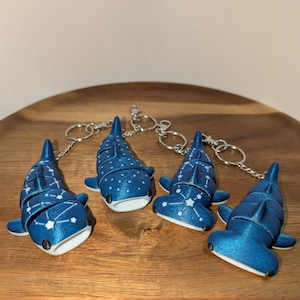 May include: Four articulated shark keychains in a metallic blue finish. Two feature white constellation patterns on their backs, while the other two have white polka dots. Each shark has a white underside, black eyes, and a silver metal keyring attached.
