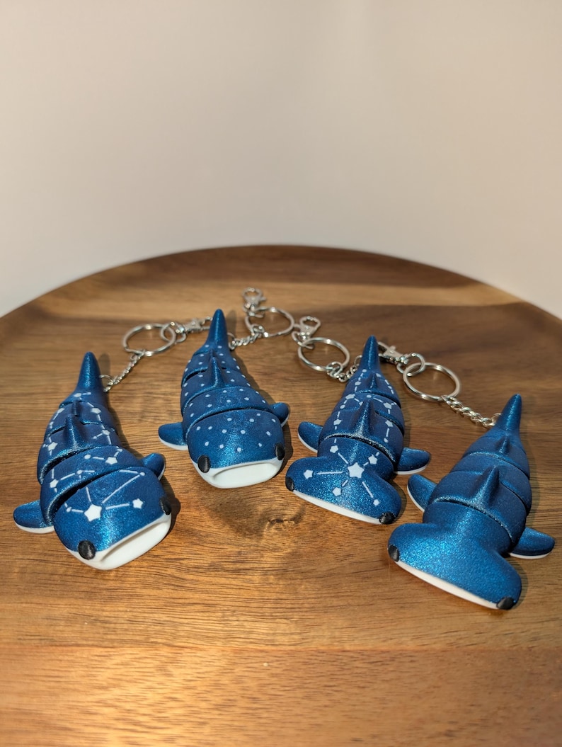 May include: Four articulated shark keychains in a metallic blue finish. Two feature white constellation patterns on their backs, while the other two have white polka dots. Each shark has a white underside, black eyes, and a silver metal keyring attached.