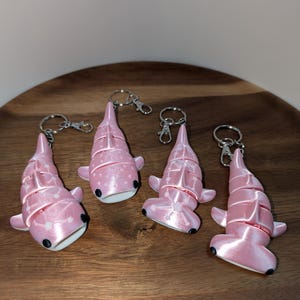 May include: Four articulated shark keychains in a light pink colour. Two keychains feature a whale shark design with white dot patterns, and two feature a hammerhead shark design. Each shark has small black eyes and a silver metal keyring attached.