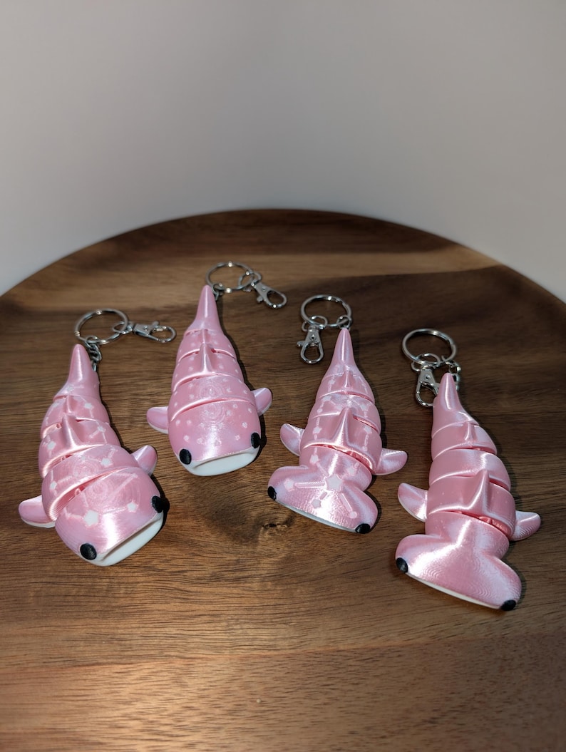 May include: Four articulated shark keychains in a light pink colour. Two keychains feature a whale shark design with white dot patterns, and two feature a hammerhead shark design. Each shark has small black eyes and a silver metal keyring attached.