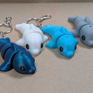 May include: Four articulated dolphin keychains in metallic blue, solid white, bright cyan, and matte gray. Each dolphin features a segmented body, a dorsal fin, and two side fins, with black circular eyes and a small hole on the top of the head.