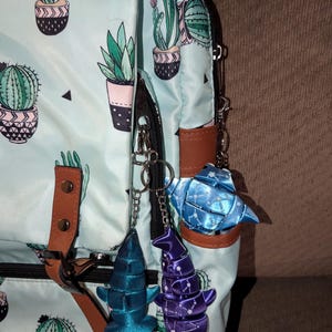 May include: A light blue backpack featuring a repeating cactus print in green and pink pots, accented with brown faux leather straps. Three ribbon-based charms in blue, purple, and light blue are attached to the black zipper hardware.