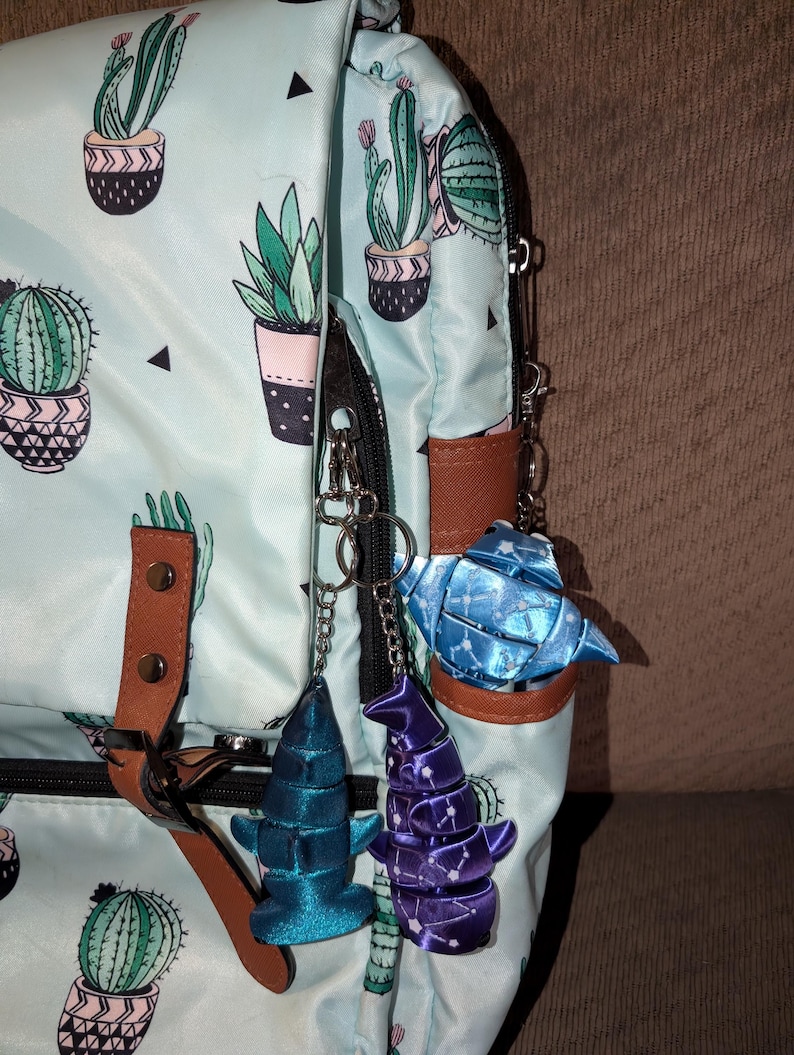 May include: A light blue backpack featuring a repeating cactus print in green and pink pots, accented with brown faux leather straps. Three ribbon-based charms in blue, purple, and light blue are attached to the black zipper hardware.