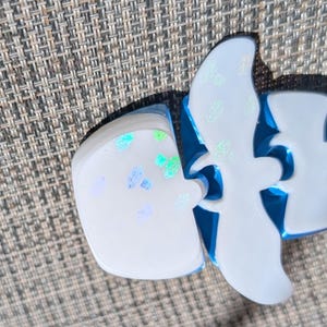 May include: A white resin object with a smooth surface featuring iridescent blue and green flecks. The object has a distinct bird-like silhouette with curved wings and a rounded base, showcasing a glossy finish and a bright blue interior edge.