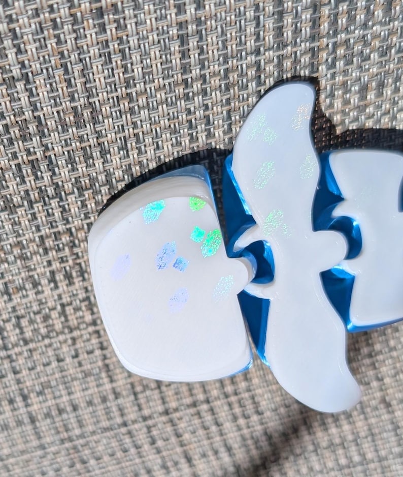 May include: A white resin object with a smooth surface featuring iridescent blue and green flecks. The object has a distinct bird-like silhouette with curved wings and a rounded base, showcasing a glossy finish and a bright blue interior edge.
