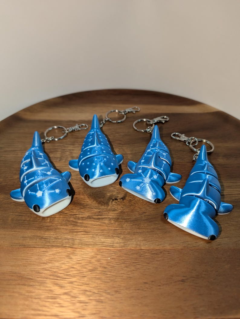 May include: Four articulated shark keychains in a metallic blue finish with white constellation patterns. Two keychains are whale sharks and two are hammerhead sharks, each featuring a silver-toned metal split ring and a lobster claw clasp.