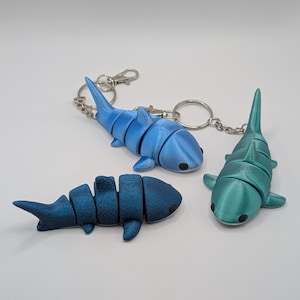 May include: Three articulated shark keychains in shades of blue and teal. The sharks have segmented bodies and small black eyes. Two keychains are attached to silver keyrings. The sharks are displayed on a white background.