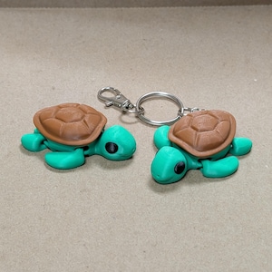May include: Two turtle keychains. Each keychain features a green turtle body with a brown shell. The turtles are attached to a silver-colored keyring and clasp. The keychains are displayed on a light brown surface.