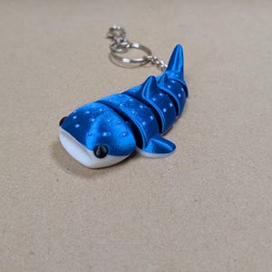 May include: A blue and white articulated whale shark keyring featuring a segmented body, a flat white underside, and small white spots across the top. The shark is attached to a silver-coloured metal split ring and a short chain for keys.